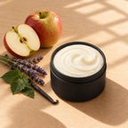 Apple Lavender and Patchouli whipped tallow body butter in an open matte black tin beside a fresh apple, lavender sprigs, patchouli leaves and a vanilla bean