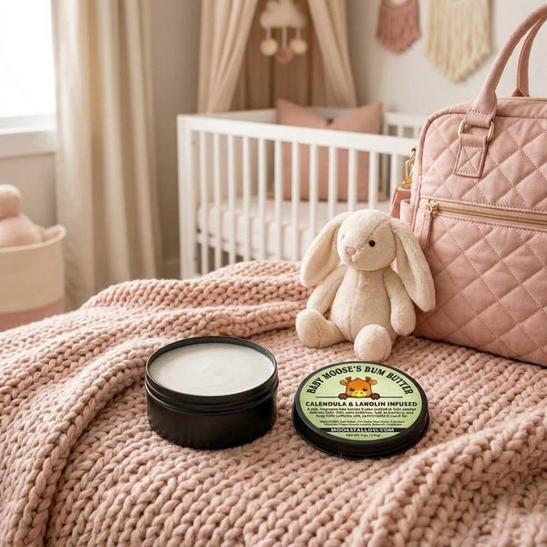 Baby Bum Butter natural tallow and zinc oxide diaper cream — open and closed tins with the Baby Moose's Bum Butter calendula and lanolin label on a blush-pink knit blanket in a baby girl's nursery